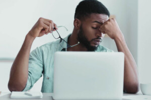 A young Black man rubbing his eyes while working on the laptop & holding glasses. Representing how stress in your 20's can be overwhelming. Get started with a young adult therapist in Medford, NJ today.
