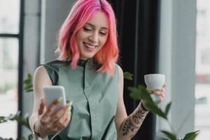 A happy woman with bright pink hair smiling while holding her phone & cup. Therapy for young adults in Medford, NJ can help teens & adults tackle anxiety, life changes & more. 
