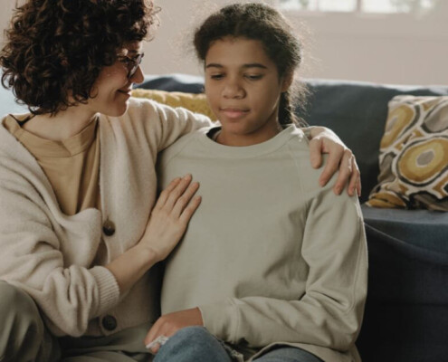 A woman gently comforts a young girl on a couch, with a supportive arm around her in a cozy living room