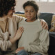 A woman gently comforts a young girl on a couch, with a supportive arm around her in a cozy living room