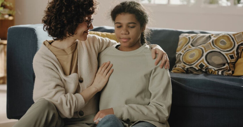 A woman gently comforts a young girl on a couch, with a supportive arm around her in a cozy living room