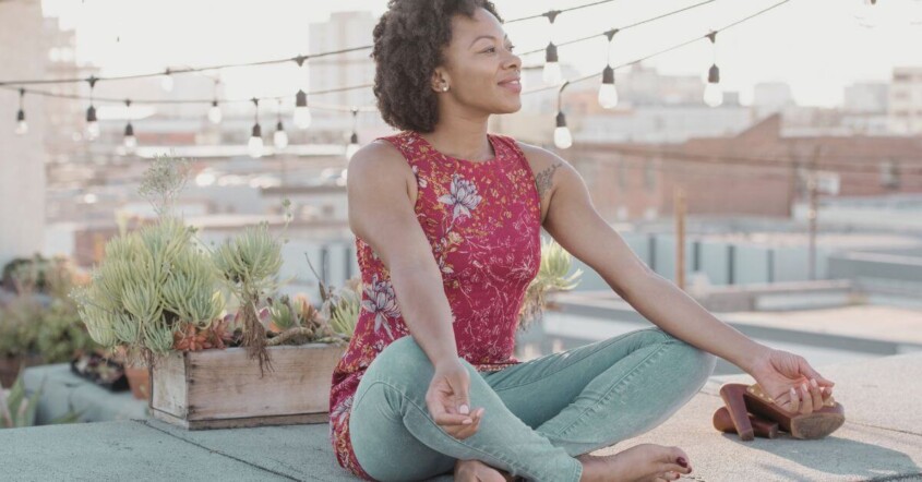 A young adult woman meditates peacefully on a rooftop at sunset.