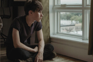 A young person sits on the floor, gazing thoughtfully out a window.