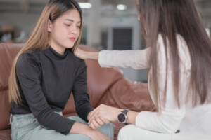 A female therapist comforts a young woman by holding her hand and touching her shoulder.