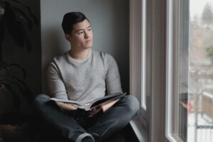 A young adult sits by a window reading a book and looking outside thoughtfully.