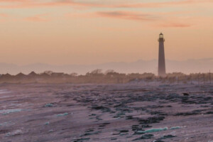 A New Jersey lighthouse stands near the shore at sunset with a misty beach in the foreground.