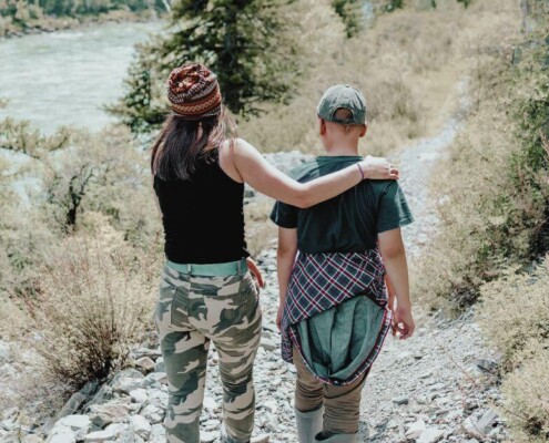 Mom and teen walk side by side on a forest trail, one with an arm around the other.
