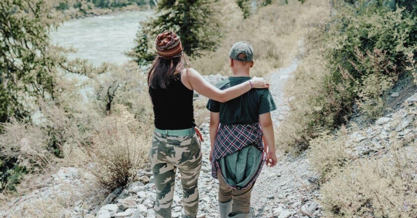 Mom and teen walk side by side on a forest trail, one with an arm around the other.