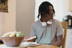 A teen wearing headphones sits at a table eating toast, looking to the side.