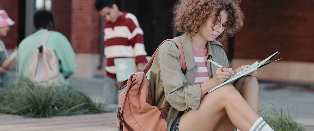 A student sits outside writing in a notebook while others stand nearby.