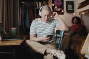 A student wearing headphones looks at their phone in a dorm room, while another sits on a bed in the background.