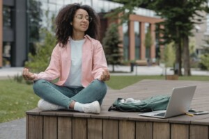 A student meditates outdoors on campus, sitting cross-legged near a laptop and backpack.