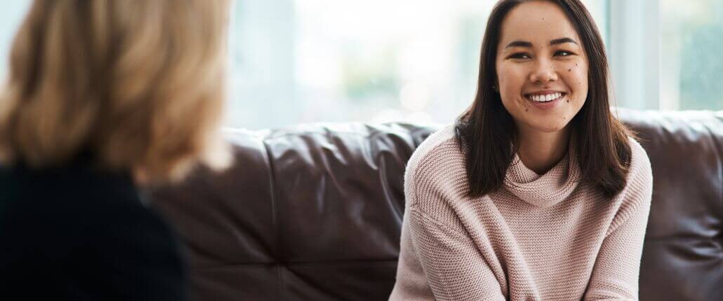 A smiling Asian college student sits on a couch, talking with a therapist.