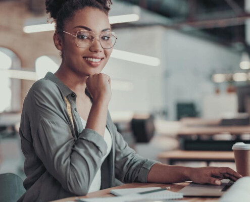 A smiling young woman with glasses sits at a desk, working on a laptop in a modern office.