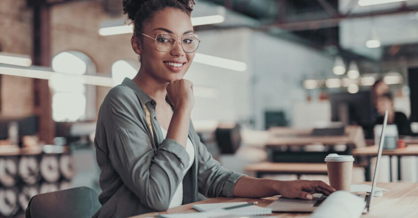 A smiling young woman with glasses sits at a desk, working on a laptop in a modern office.
