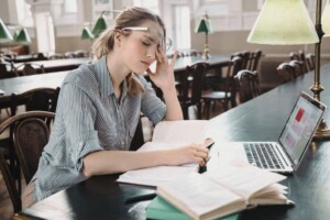 Tired young college student studying in a library with books and a laptop.