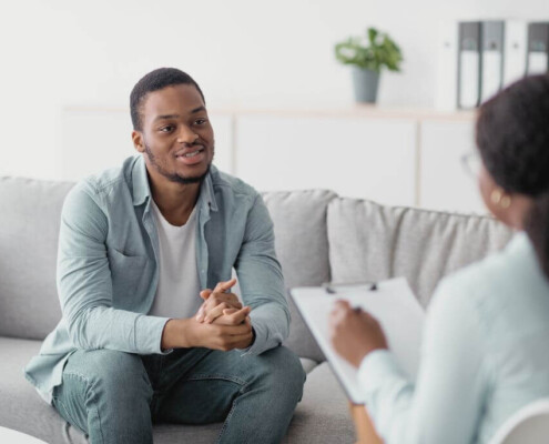 A man sits on a couch talking with a therapist, who is holding a clipboard and taking notes.