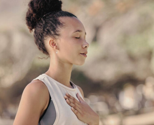 A young woman stands outdoors with her eyes closed, one hand on her chest, practicing calm, mindful breathing.