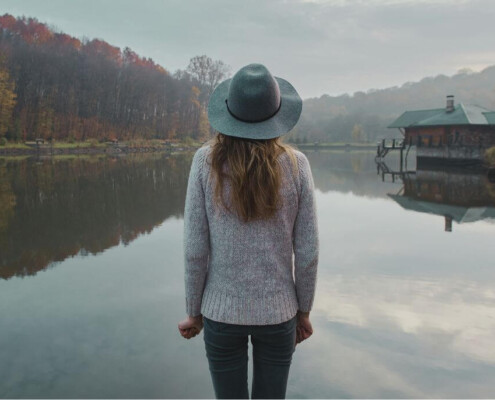 A woman wearing a hat stands at the edge of a still lake, looking out at the water and the autumn trees and cabin across the shore.