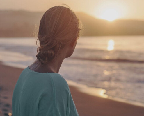 A young woman stands on a beach at sunset, looking out toward the ocean. If you're trying to balance your career, school, & dating, discover how young adult therapy in Cherry Hill, NJ can help you navigate it all.