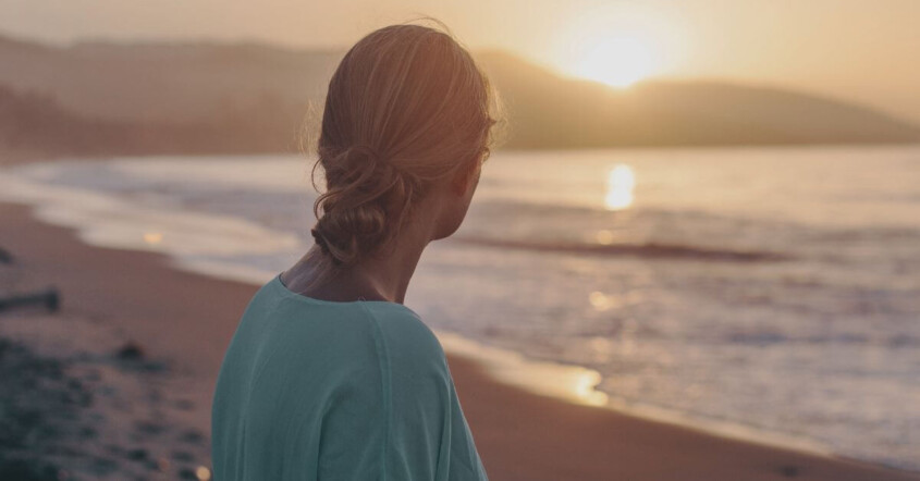 A young woman stands on a beach at sunset, looking out toward the ocean. If you're trying to balance your career, school, & dating, discover how young adult therapy in Cherry Hill, NJ can help you navigate it all.
