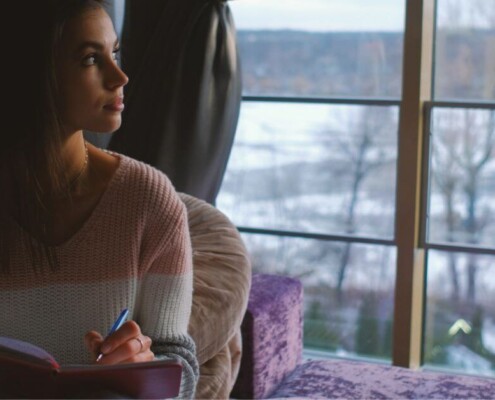 A young woman sits indoors by a large window on a winter day, journaling in a notebook. Discover how young adult therapy in Medford, NJ can help you build confidence. Read more here!