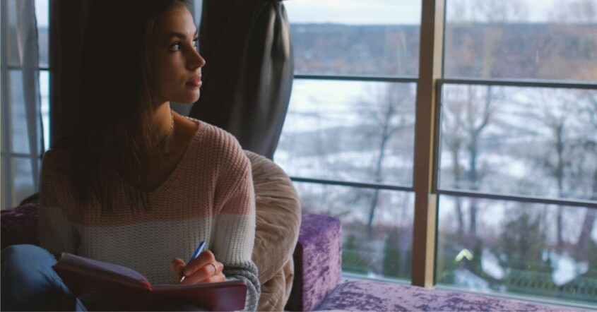 A young woman sits indoors by a large window on a winter day, journaling in a notebook. Discover how young adult therapy in Medford, NJ can help you build confidence. Read more here!