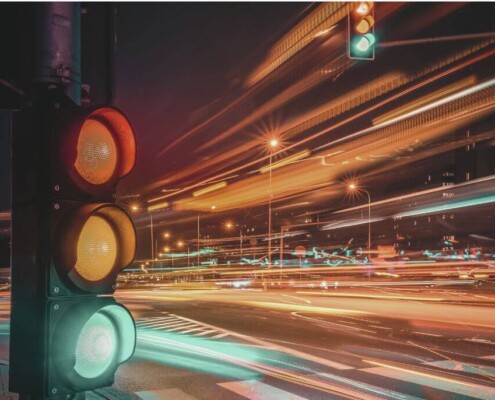 A traffic light glows red and yellow at a busy intersection at night, with long streaks of colorful light trails from passing cars creating a sense of fast motion