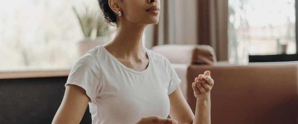 A woman sits indoors with her eyes closed, holding her hands in a relaxed, meditative position as she practices deep breathing or mindfulness.