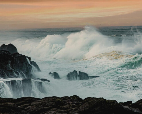 Large ocean waves crash against dark, rocky cliffs under a soft orange sky at sunset.