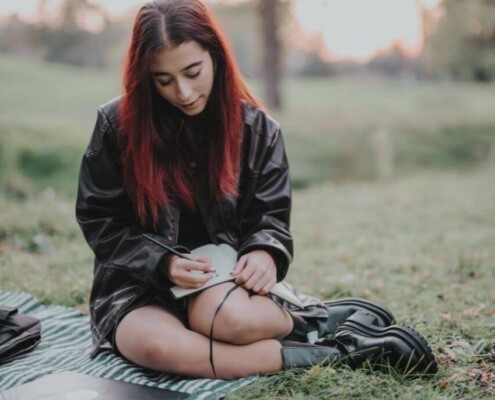 A young woman with red hair sits on a blanket outdoors, journaling in a notebook with a pen in her hand. If you're struggling to find yourself, discover what it means by reading our blog. Our young adult therapists in Medford, NJ can help you.