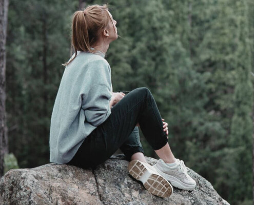 A woman sits on a large rock in the woods, looking up toward the sky while surrounded by tall trees. A young adult therapist in New Jersey can help you navigate adulthood with confidence. Learn more here!