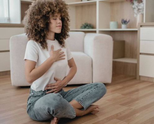 A woman sits cross-legged on the floor with her eyes closed, one hand on her chest and the other on her abdomen, practicing deep breathing or mindfulness. A therapist for young adults in New Jersey can help you find your identity. Reach out today to learn how to get started. 