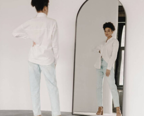 A woman stands in front of a tall mirror, looking at her reflection while resting one hand on her hip. Young adult therapy in Medford, NJ is here to support individuals with life transitions. Discover more by reading our blog.