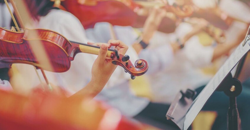 Close-up of musicians playing violins in an orchestra, with hands on the strings and bows moving in unison.