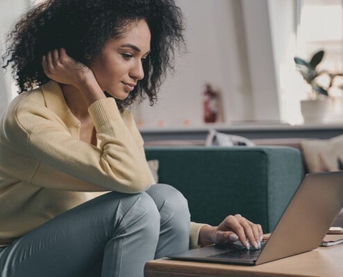 ○ A woman sits on a couch working on her laptop, leaning forward with a focused expression in a bright, cozy living room.