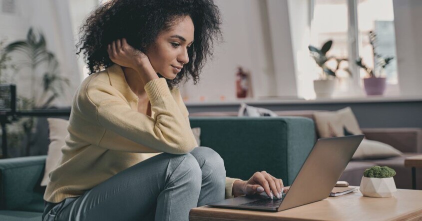 ○ A woman sits on a couch working on her laptop, leaning forward with a focused expression in a bright, cozy living room.