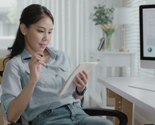A woman sits at her desk reviewing information on a tablet, with charts and graphs displayed on a computer screen beside her.