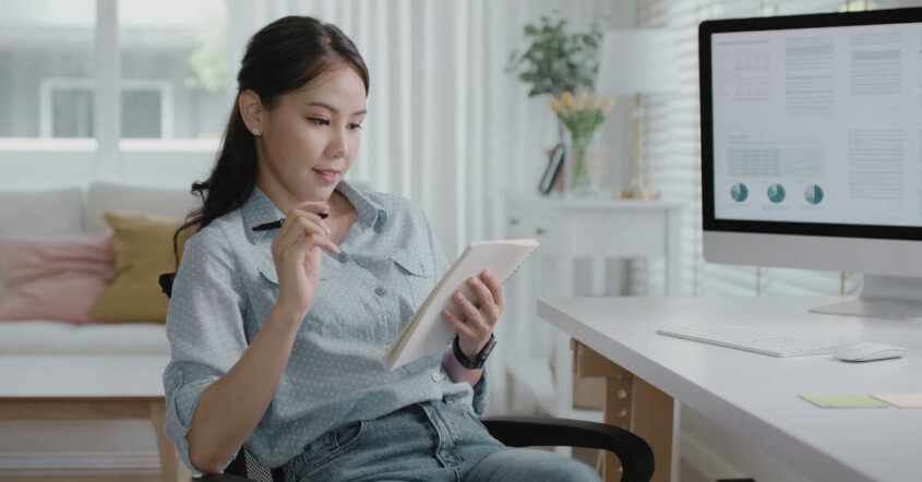 A woman sits at her desk reviewing information on a tablet, with charts and graphs displayed on a computer screen beside her.