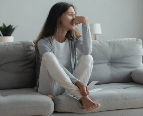 A woman sits curled up on a couch, holding a piece of paper and crying with her hand covering part of her face.