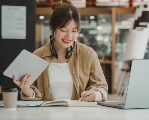A woman smiles while studying at a desk, holding a tablet in one hand and writing in a notebook with a laptop and books nearby