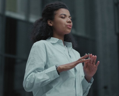 A woman stands outdoors with her eyes closed, taking a deep breath while holding her hands in a relaxed, grounding position.