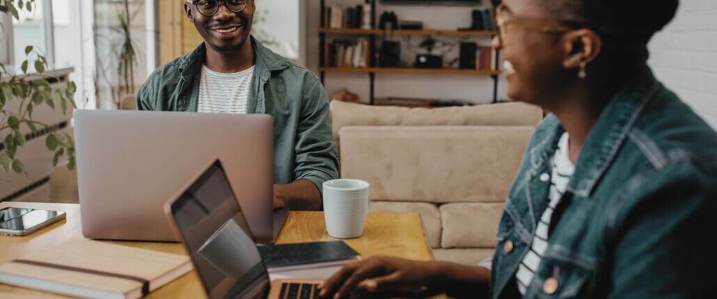 Two people sit across from each other at a table, smiling and working on their laptops in a cozy, modern workspace.