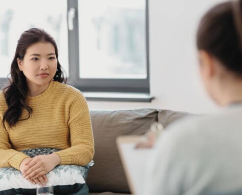 A woman sits on a couch holding a pillow in her lap, listening thoughtfully to a therapist who is taking notes.