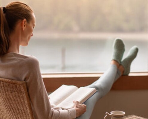 Woman reading a book while resting her feet on a windowsill. Representing how beginning therapy in January can help you reach your goals in a supportive way. Contact us today to start healing.