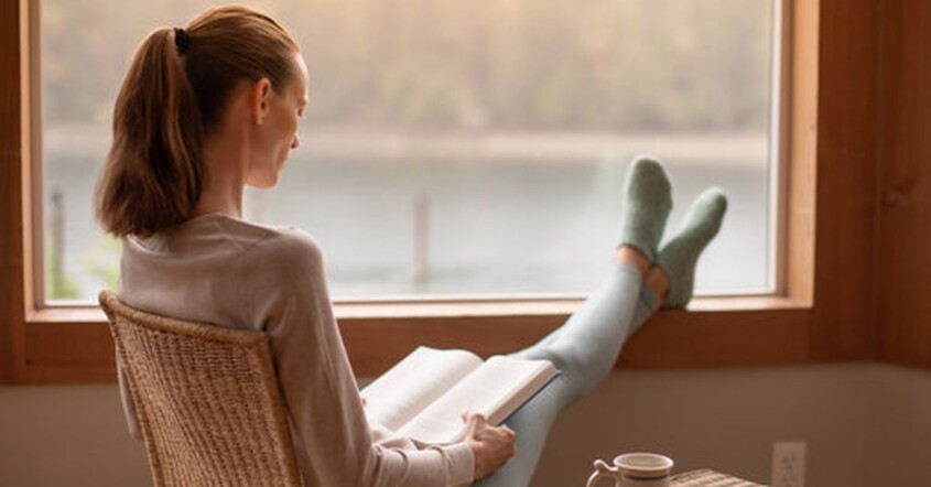 Woman reading a book while resting her feet on a windowsill. Representing how beginning therapy in January can help you reach your goals in a supportive way. Contact us today to start healing.