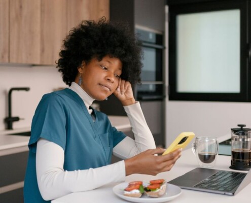 Nurse sitting at a kitchen counter during a break, looking at her phone while balancing work stress and personal responsibilities.