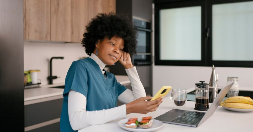 Nurse sitting at a kitchen counter during a break, looking at her phone while balancing work stress and personal responsibilities.