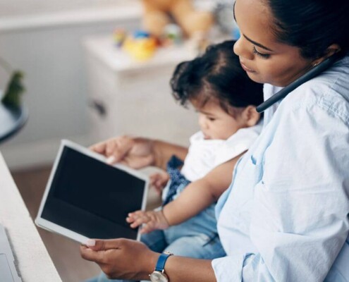 Mother working from home while holding her young child and using a phone and tablet.