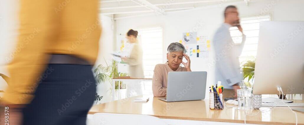 Overwhelmed older professional woman sitting at a desk in a busy office, experiencing workplace stress